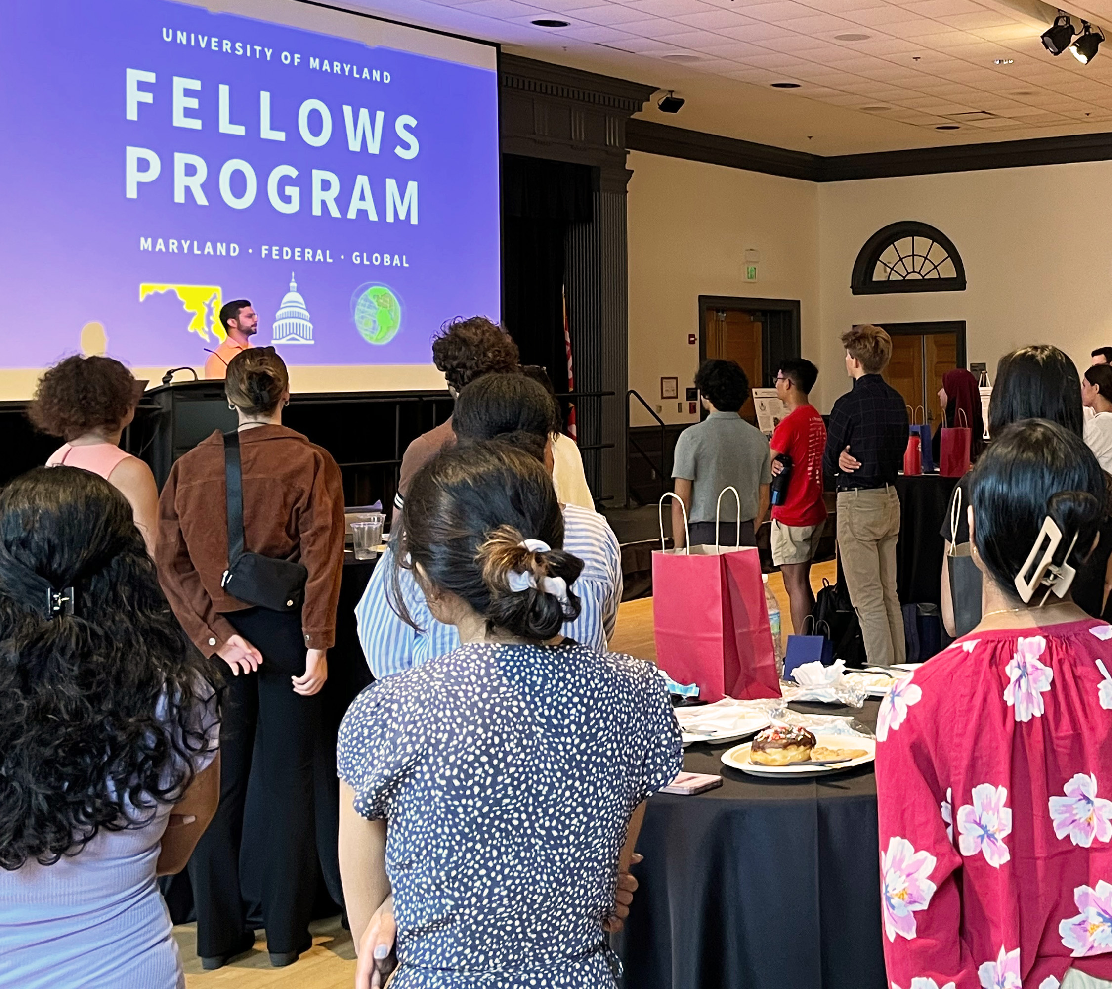 Ballroom full of students standing and listening to a man speak in front of the UMD Fellows Program logo